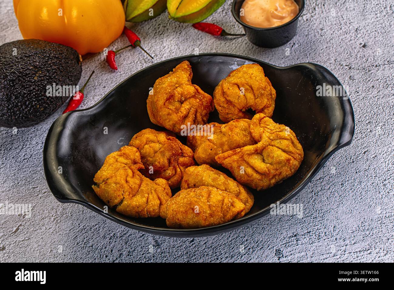 Goldene gebratene Knödel in schwarzer Keramikschale. Zu den umliegenden Zutaten gehören ganze Avocado gelbe Paprika rote Chilischoten, cremige Dippi Stockfoto