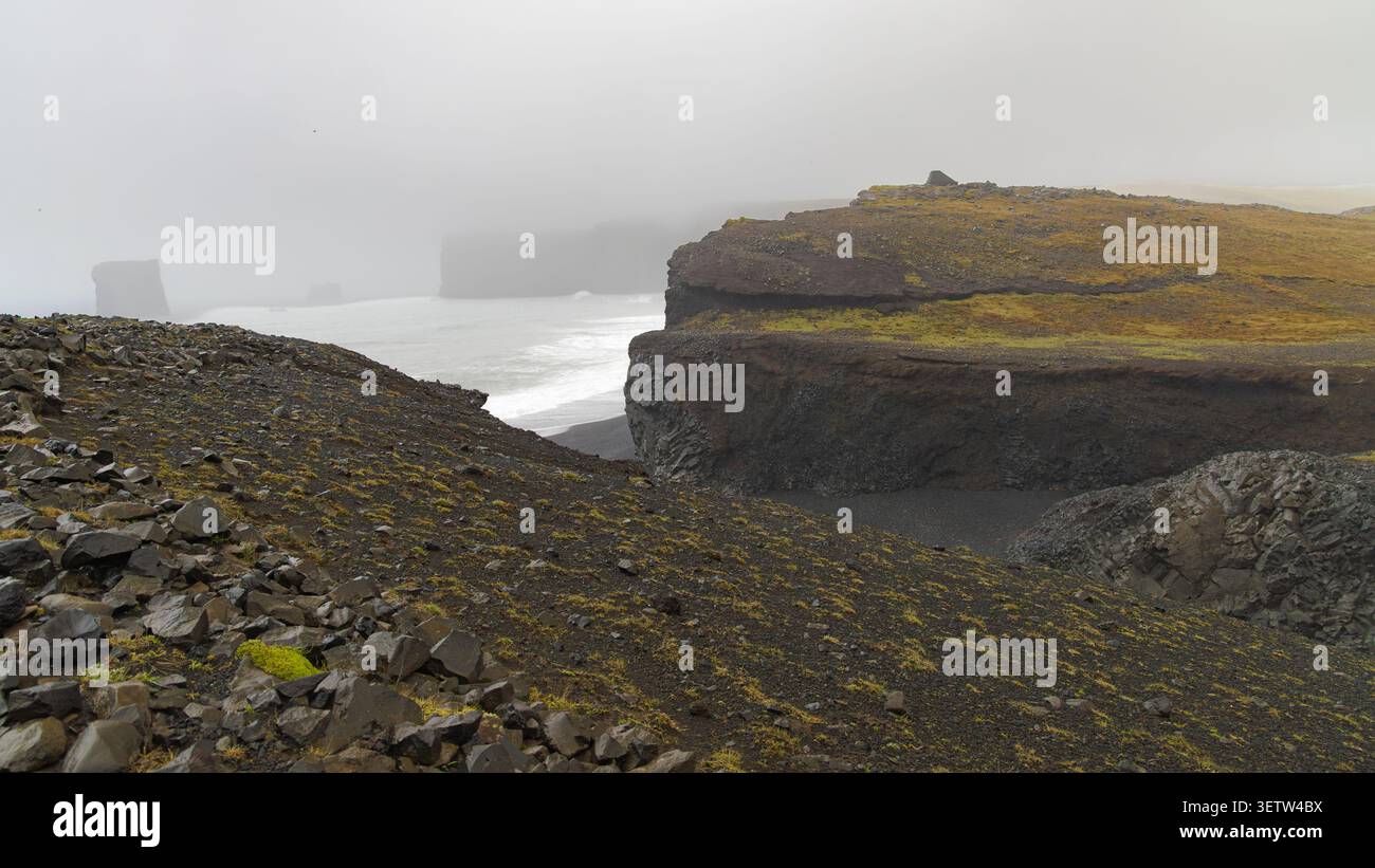 Vulkanische Klippen und felsige Landschaft von Dyrhólaey in Island mit Blick auf das Meer und den schwarzen Sandstrand unter einem nebeligen, bewölkten Himmel. Stockfoto