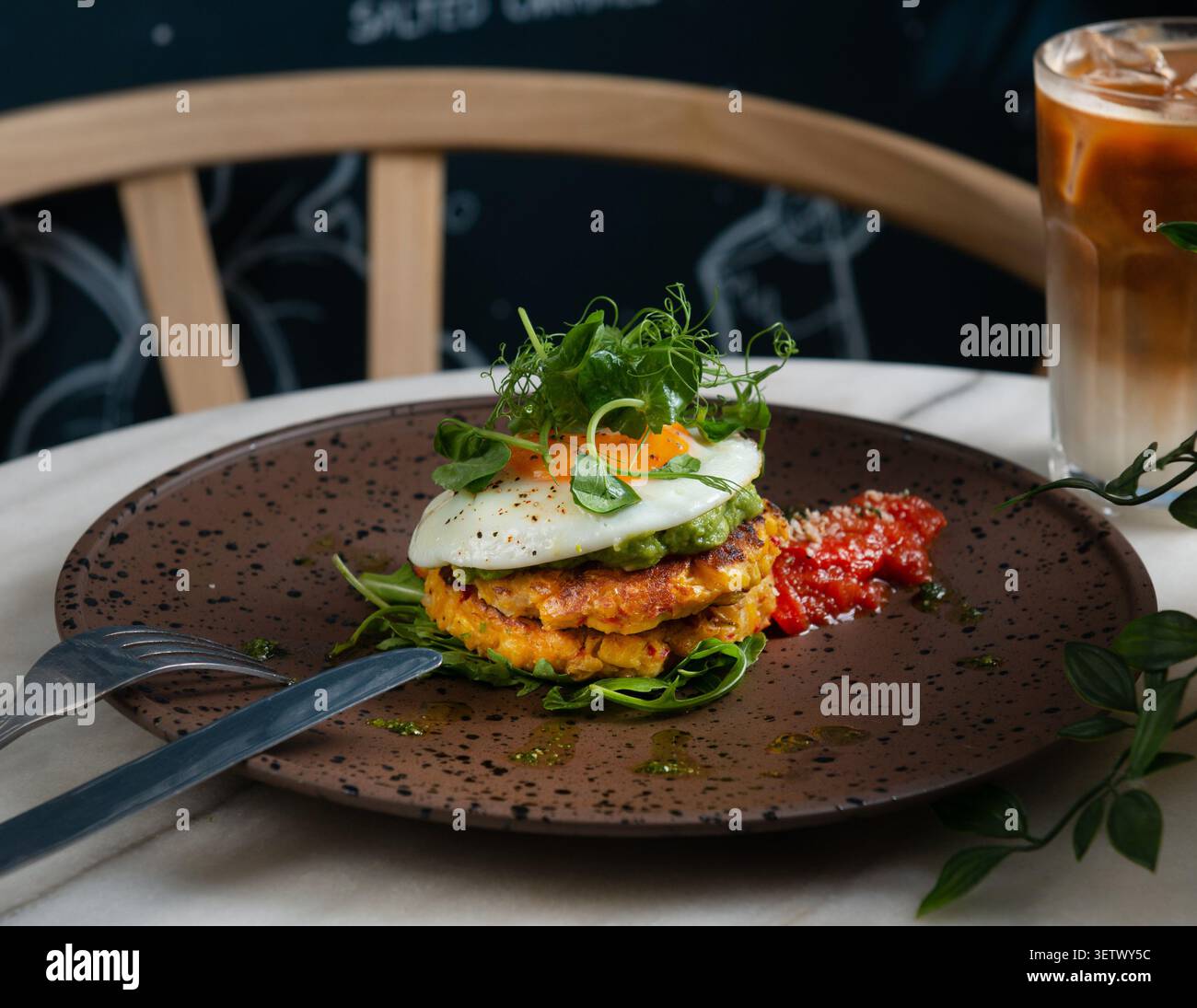 Herzhafte Maisfritter mit zerstoßener Avocado, Spiegelei und frischem Mikrogrün, serviert mit Tomaten-Relish und Eiskaffee. Stockfoto