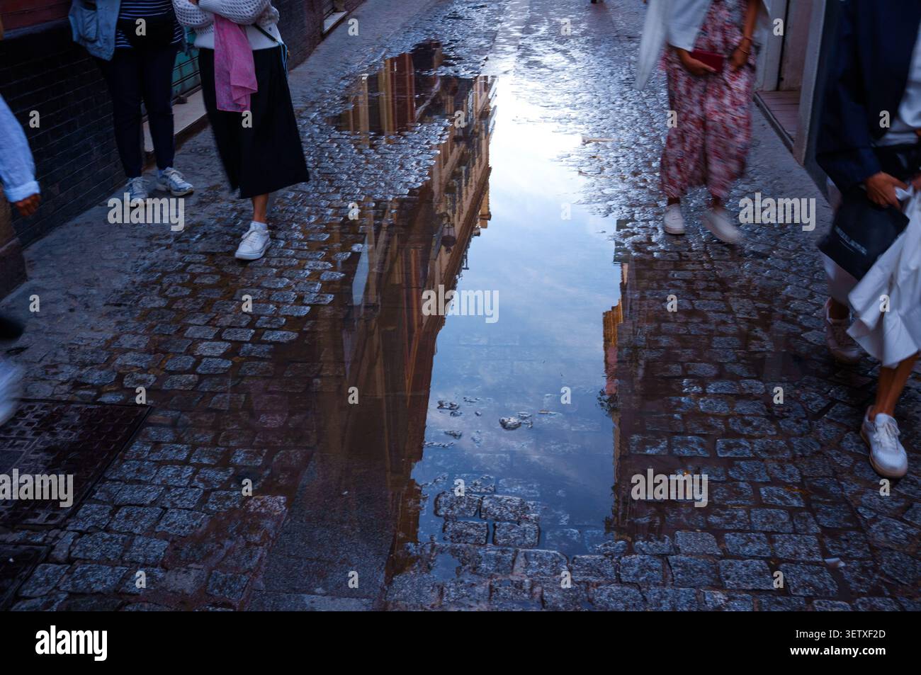 Eine Regelpfütze auf den Kopfsteinpflasterstraßen von Sevilla, Spanien, die Gebäude reflektiert, während vorbeigehende Menschen das Wasser umgehen. Stockfoto