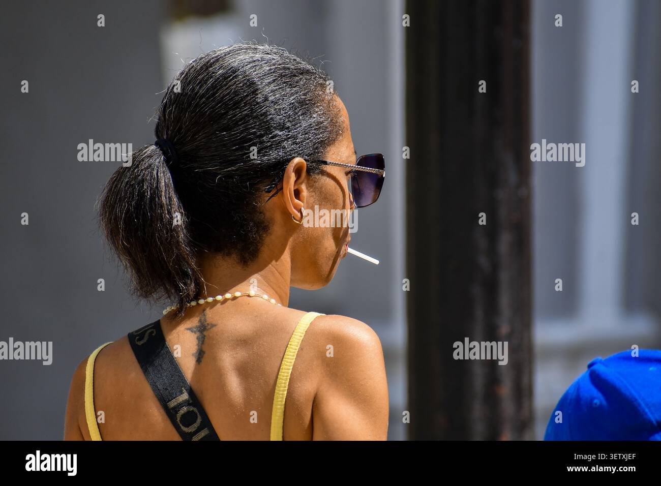 Seiten- und Rückansicht einer kubanischen Frau mit Sonnenbrille, einem Pferdeschwanz, einem kleinen Nackentattoo und einem Lutscher-Stab im Mund im Jose-Marti-Park in Ciego de Stockfoto