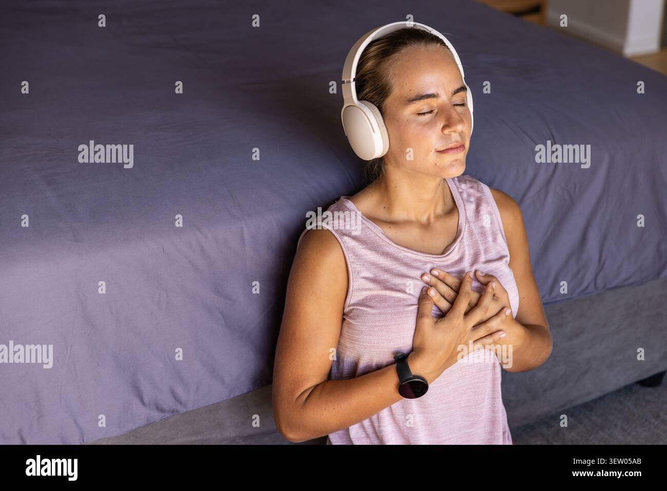Frau Mitte 20, die neben dem Bett sitzt, mit grauer Federdecke, rosa Oberseite, Kopfhörern, Smartwatch, Kopierraum Stockfoto