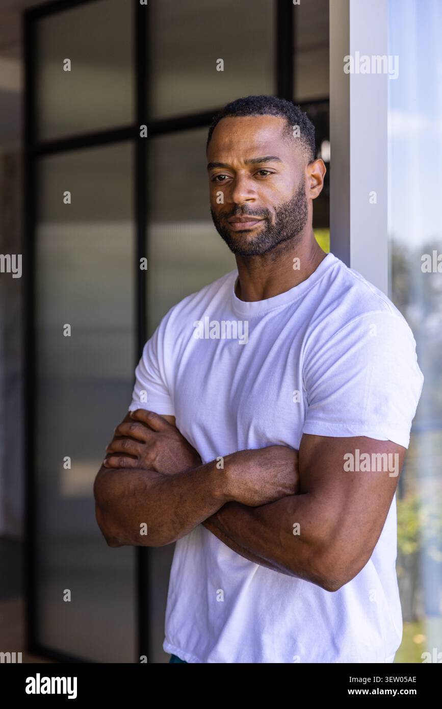 Reifer afroamerikanischer Mann, der sich auf einen Lichtrahmen im Eingang neben dunklem Glas stützt und weißes T-Stück trägt Stockfoto
