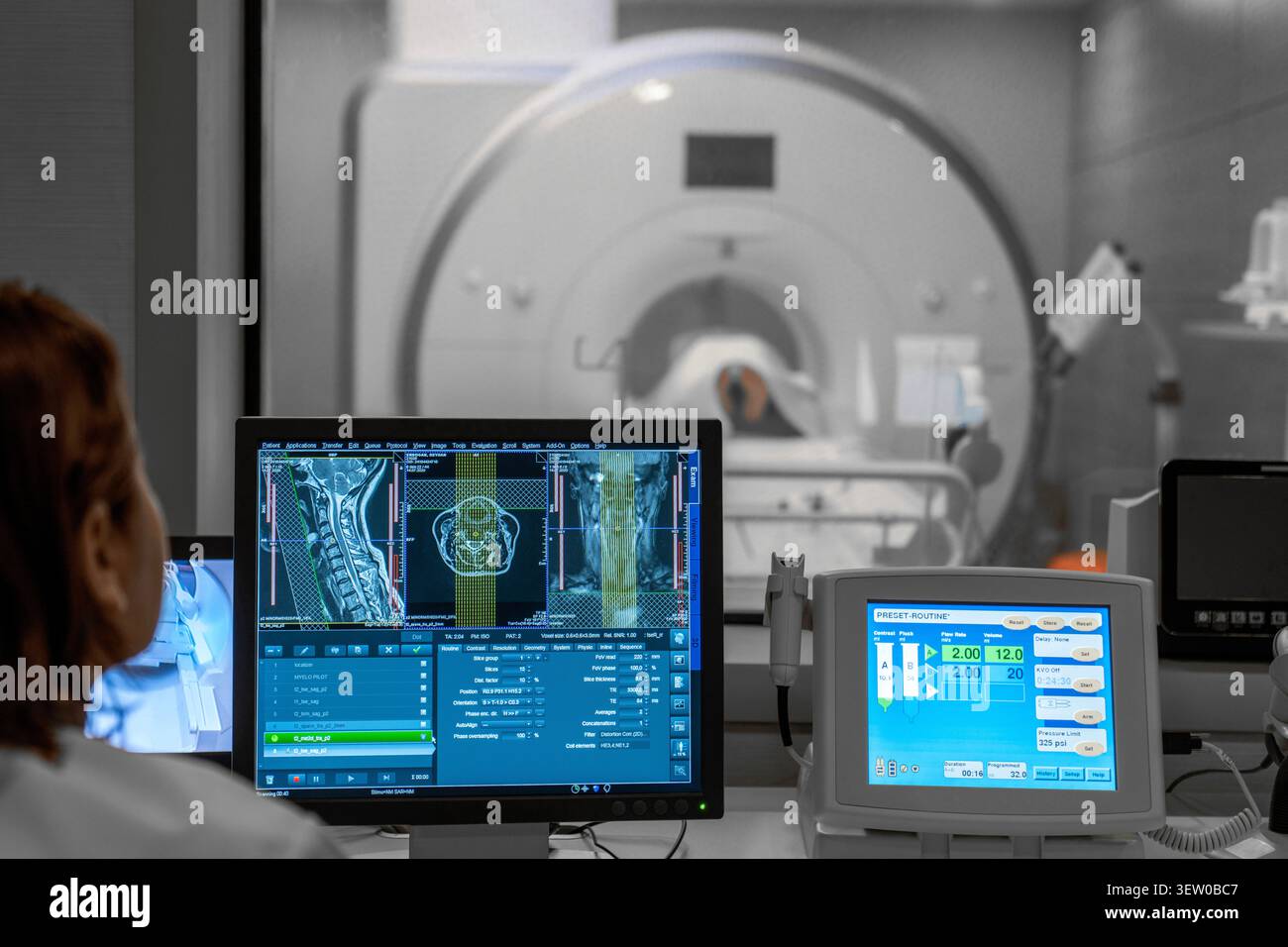 Radiologietechniker, der neben einem Patienten in einem CT- oder MRT-Scanner steht, während ein anderer Techniker die digitalen medizinischen Bilder von der Steuerung aus untersucht Stockfoto
