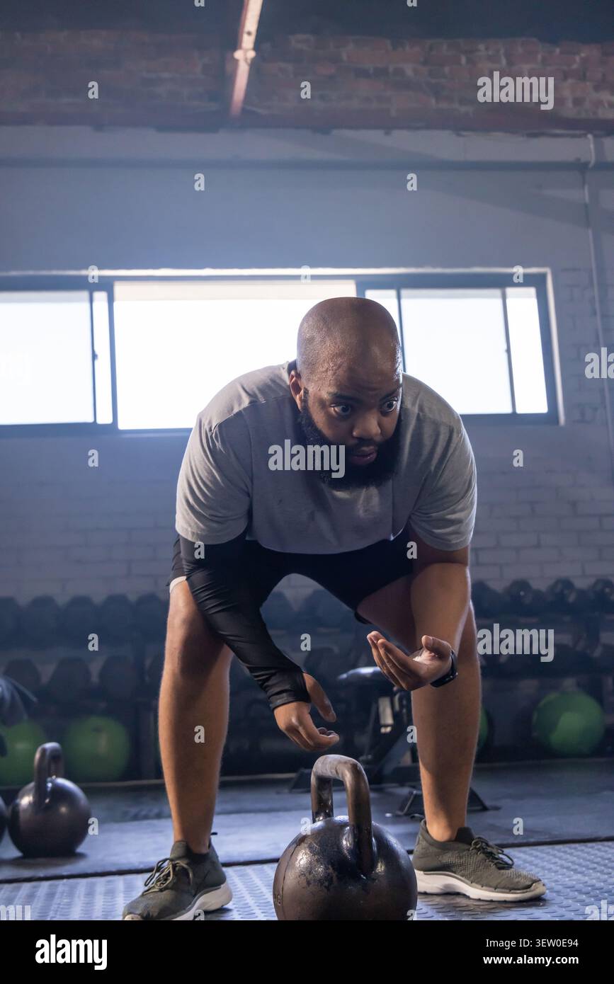 Mann, der sich darauf vorbereitet, schwere Kettlebell in einem industriellen Fitnessstudio anzuheben, mit grauem T-Shirt und schwarzen Shorts Stockfoto