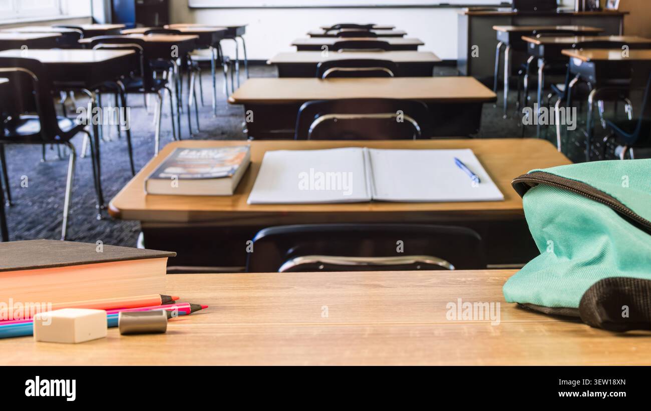 Hölzerner Klassenschreibtisch mit türkisfarbenem Rucksack, offenem Notizbuch, Buch, Farbstiften, Radiergummi und Spitzer, mit leeren Schreibtischen im Hintergrund. Stockfoto