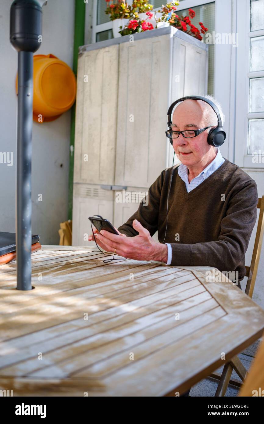 Senior Mann sitzt an einem Holztisch und hört Musik mit Kopfhörern und Smartphone Stockfoto