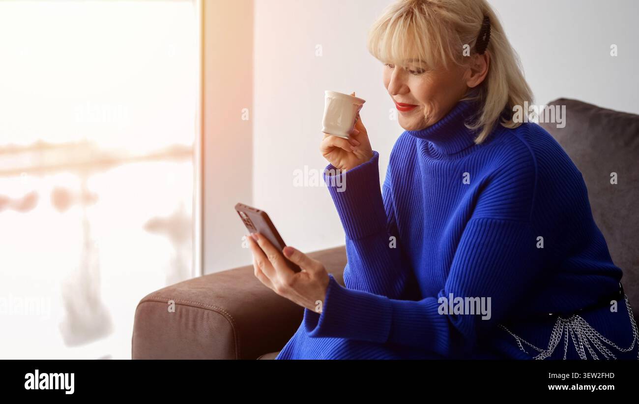 Seniorin, die sich auf einer bequemen Couch entspannt und sich glücklich mit ihrem Smartphone beschäftigt, während sie eine warme Tasse Kaffee drinnen in der Nähe eines hellen Fensters genießt Stockfoto