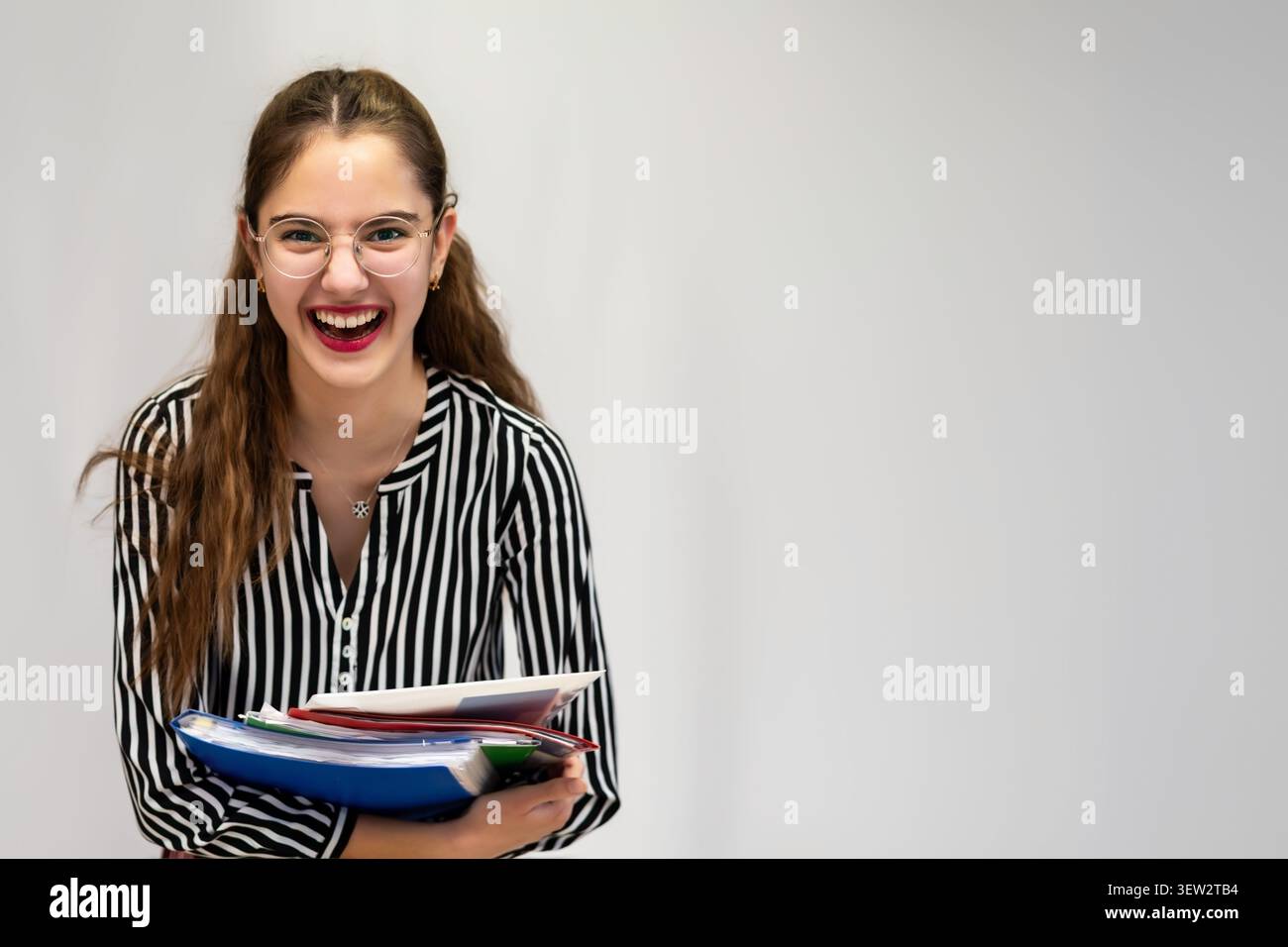 Lachende junge Frau, die im Studio Ordner hält. Fröhliche junge Frau mit Brille lacht, während sie Ordner und Papiere gegen ein hellgraues Licht hält Stockfoto