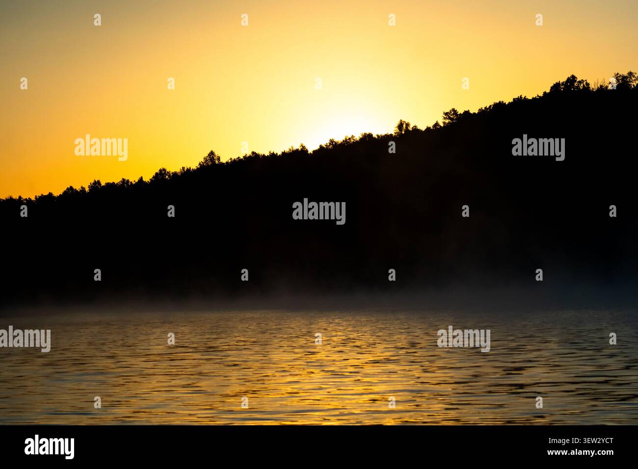 Der warme Sonnenaufgang verströmt den Kamm und verbreitet ein sanftes Leuchten über das ruhige Wasser. Nebel klebt an der Oberfläche und kleine Wellen funkeln. Die Natur begrüßt Sie am Morgen Stockfoto