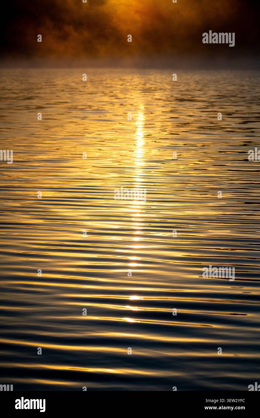 Goldene Reflektionen bilden seidenweiche Wellenmuster über den See. Lichtpunkte glitzern entlang eines hellen, vertikalen Weges. Die abstrakte Oberfläche driftet in CAL Stockfoto