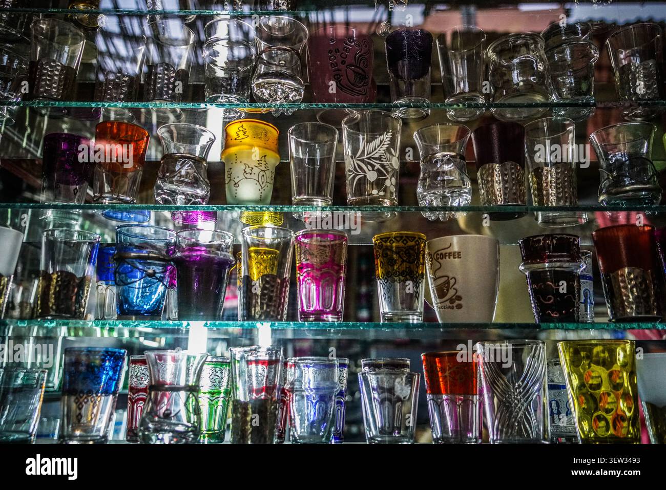 Eine Nahaufnahme auf Augenhöhe zeigt zahlreiche bunte marokkanische Teegläser und Kaffeetassen, die auf mehreren Glasregalen in einer Marktumgebung angeordnet sind. Die Stockfoto