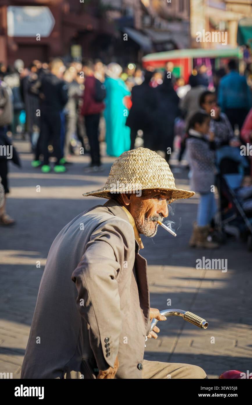 MAROKKO, MARRAKESCH - 03. Dezember 2017: Ein Mann mit Strohhut und hellbraunem Anzug sitzt mit einer Zigarette im Mund im Vordergrund, mit Blick auf das Rig Stockfoto