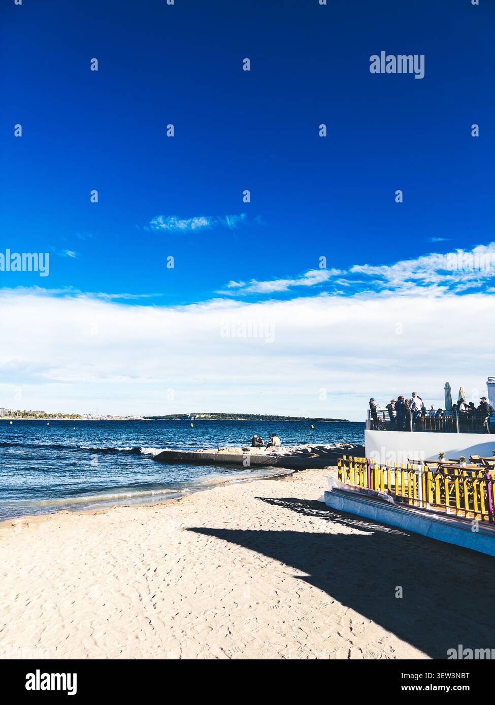 Sonnige Strandszene in Cannes an der französischen Riviera, mit einer Terrasse am Wasser und klarem blauem Himmel über dem Mittelmeer - Smartphone-aufgenommenes Stockfoto
