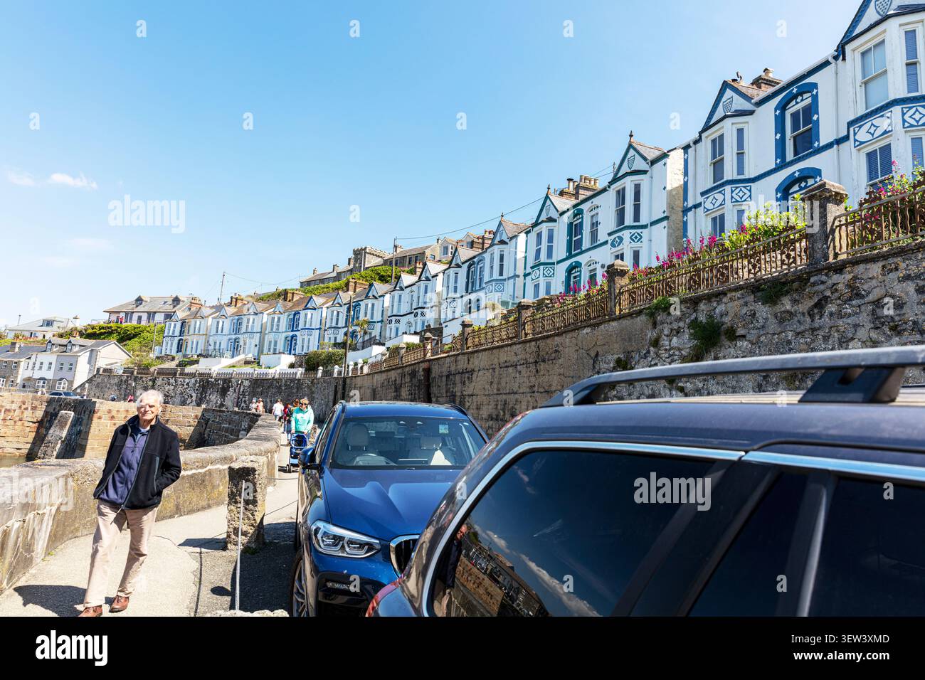 Eine Straßenszene in Porthleven, Cornwall, mit weißen und blauen Häusern am Meer, die auf einer erhöhten Steinmauer über dem Ufer errichtet wurden. Autos stehen alle Stockfoto