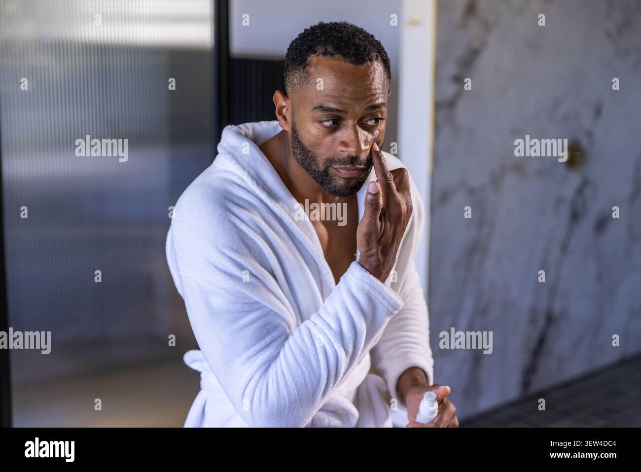 Reifer afrikanischer Mann, der in einem weißen Plüschmantel im Badezimmer sitzt und Creme aufträgt, der eine kleine Pumpflasche hält Stockfoto