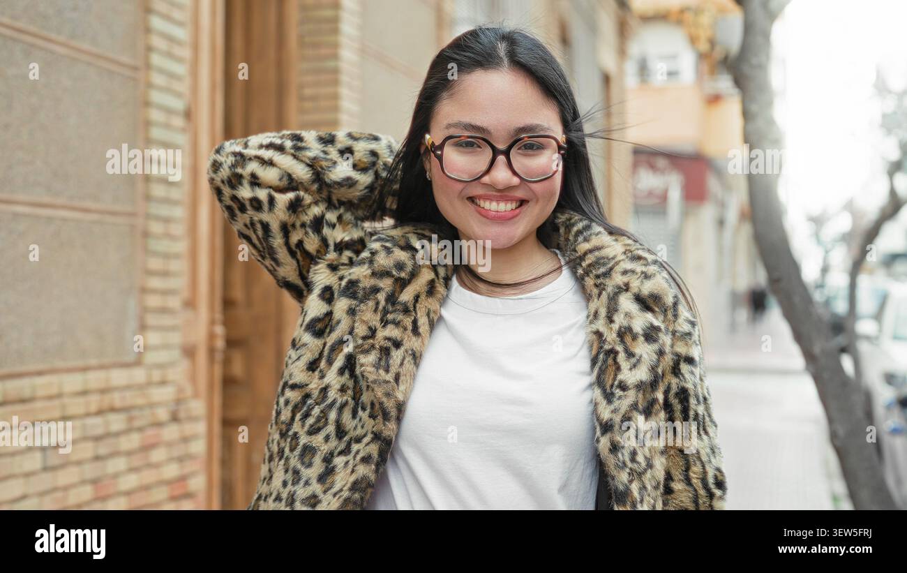 Frau mit Brille und Leopardenpelz lächelt mit der Hand hinter dem Kopf auf der Straße und lehnt sich an ein Gebäude; ungezwungenes Vertrauen. Stockfoto