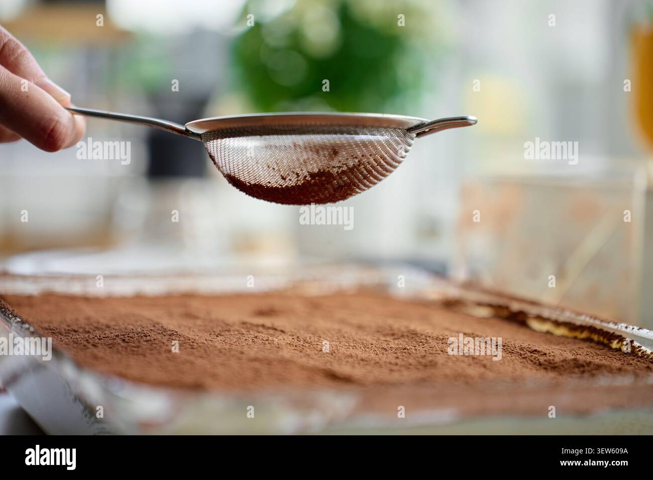 Die Hand hält das Sieb über dem mit Kakaopulver bedeckten Tiramisu-Dessert in einer Glasschale. Konzept des hausgemachten Desserts Stockfoto
