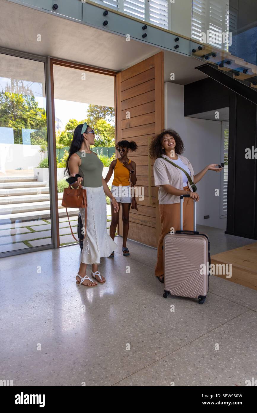 Verschiedene Freundinnen betreten das moderne Foyer durch eine große Holztür und halten einen roségoldenen Koffer Stockfoto