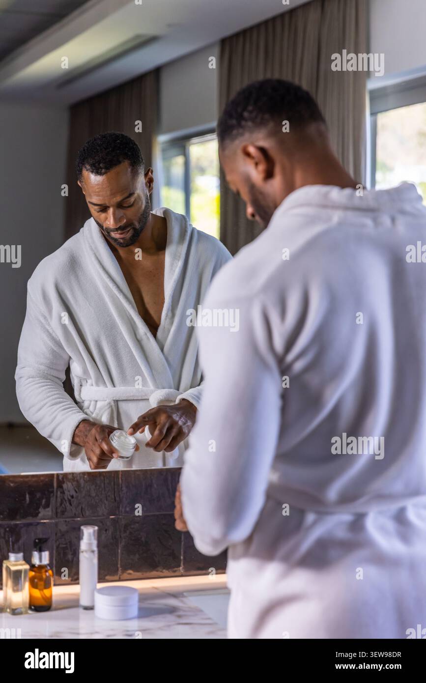Afroamerikaner mit weißem Bademantel, der Creme aus weißem Glas am Badezimmertisch aufträgt Stockfoto