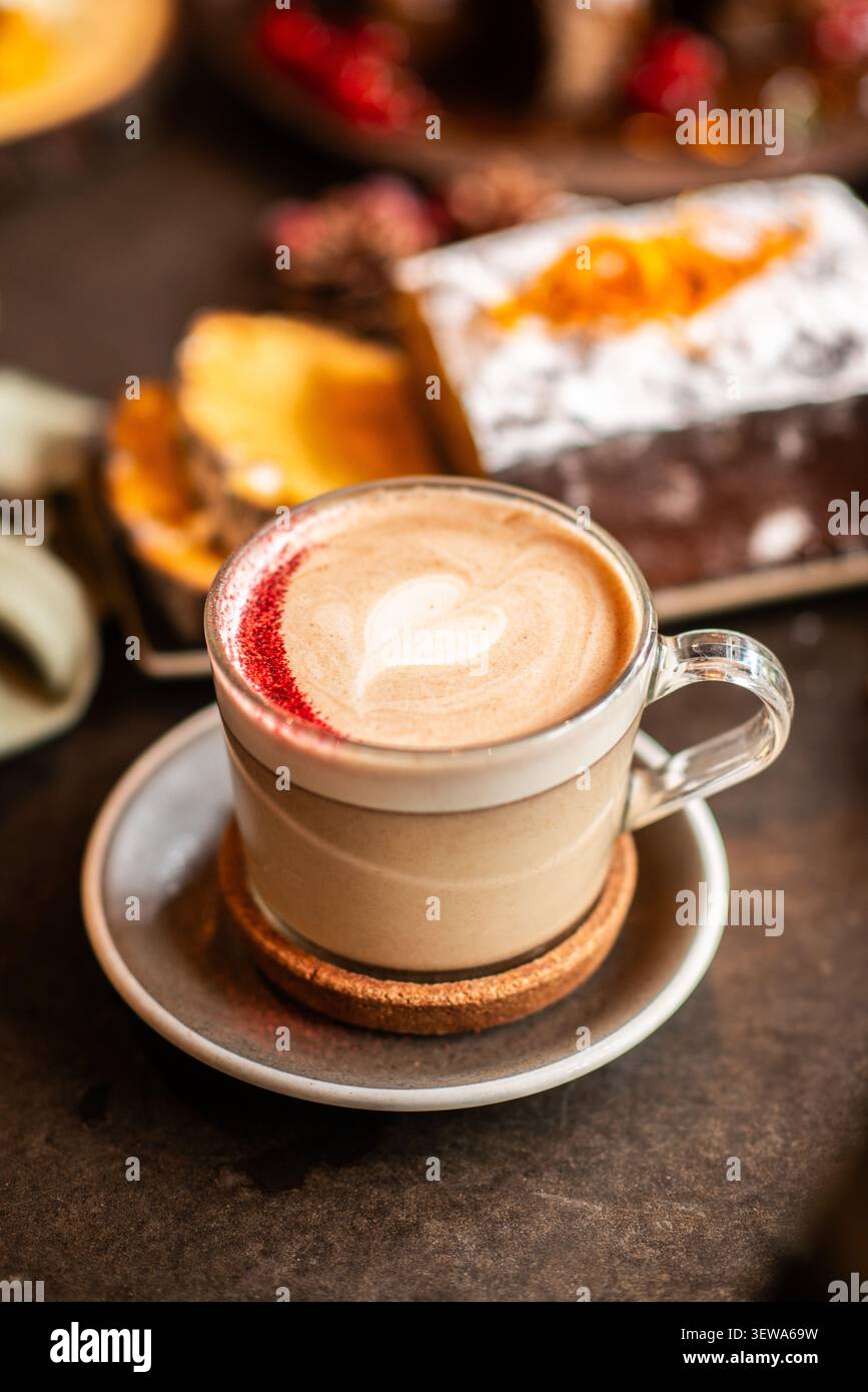 Eine Glasschale Cappuccino mit herzförmiger Latte Art und rotem Puderstaub, serviert mit geschnittenem Urlaubskuchen im Hintergrund. Stockfoto