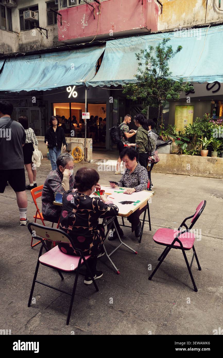 Lokale ältere Frauen spielen Kartenspiele an einem Klapptisch im Freien in einer Gasse in der Nähe eines modernen Cafés in Hongkong. Stockfoto