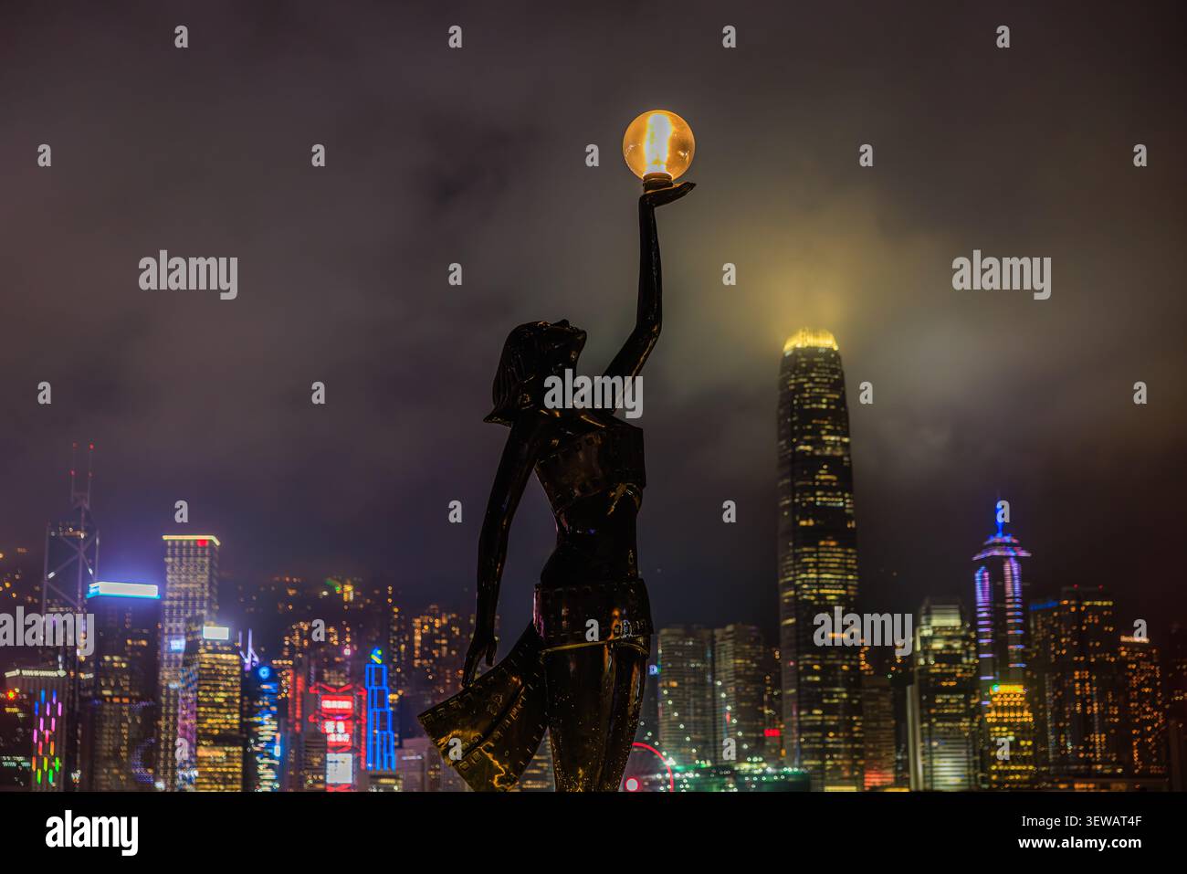 Ein dramatischer Nachtblick auf eine Bronzestatue, die eine leuchtende Kugel über der farbenfrohen beleuchteten Skyline von Hongkong in einer stimmungsvollen filmischen Stadtszene hält Stockfoto