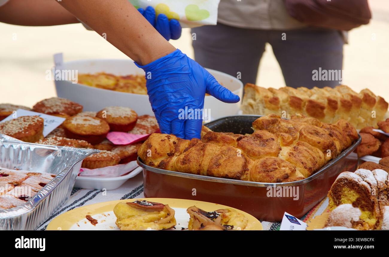Individual mit blauen Handschuhen wählt Backwaren von einem Marktstand aus, gefüllt mit verschiedenen Backwaren, Brot und Desserts auf Tischen Stockfoto