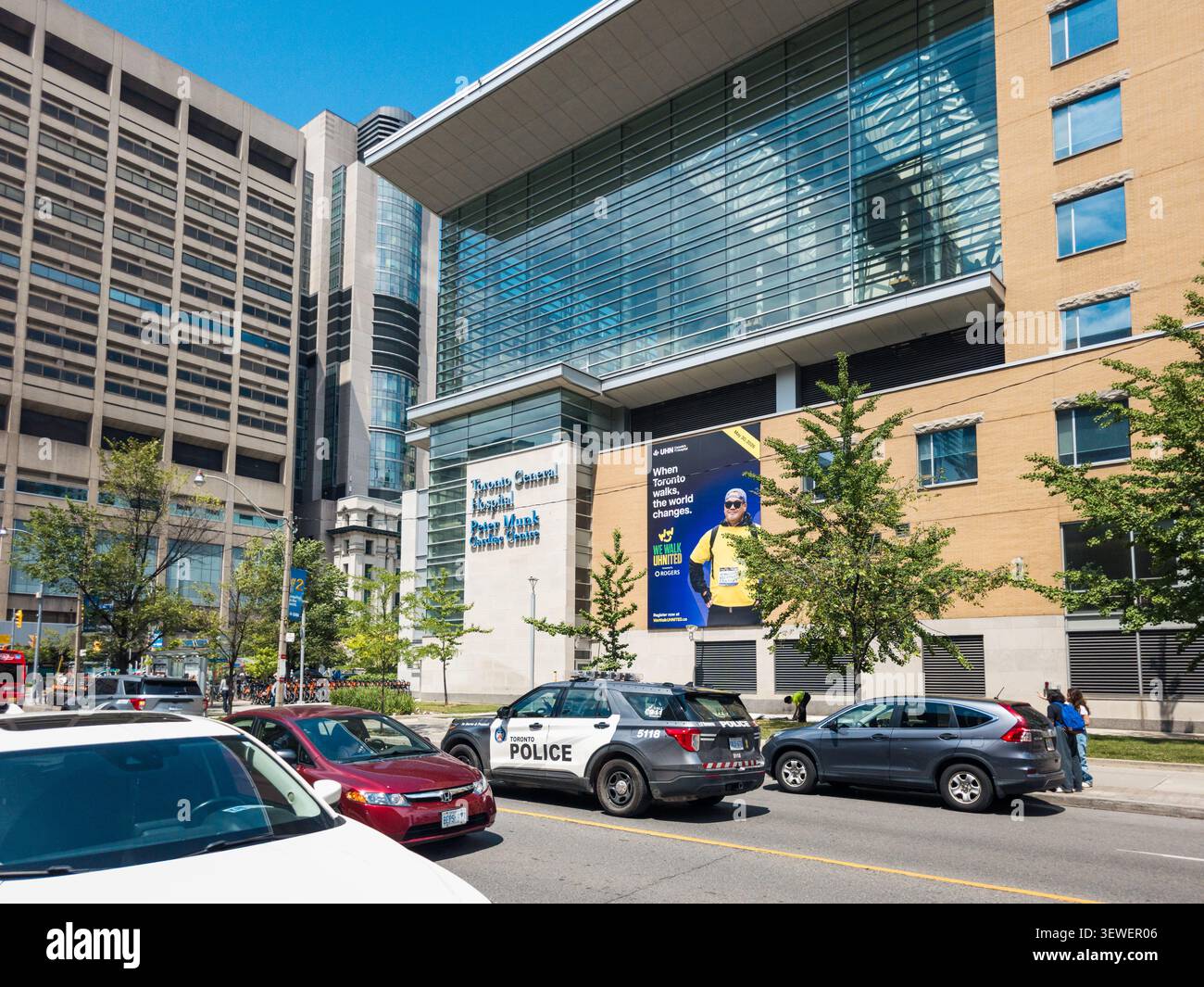 Eine Tagesansicht der Straße des Peter-Munk-Herz-Zentrums des Toronto General Hospital, das mit seiner modernen Glasfassade, einem gemeindefokussierten Banner und vorbeifahrenden Ve - Smartphone-aufgenommenes Stockfoto
