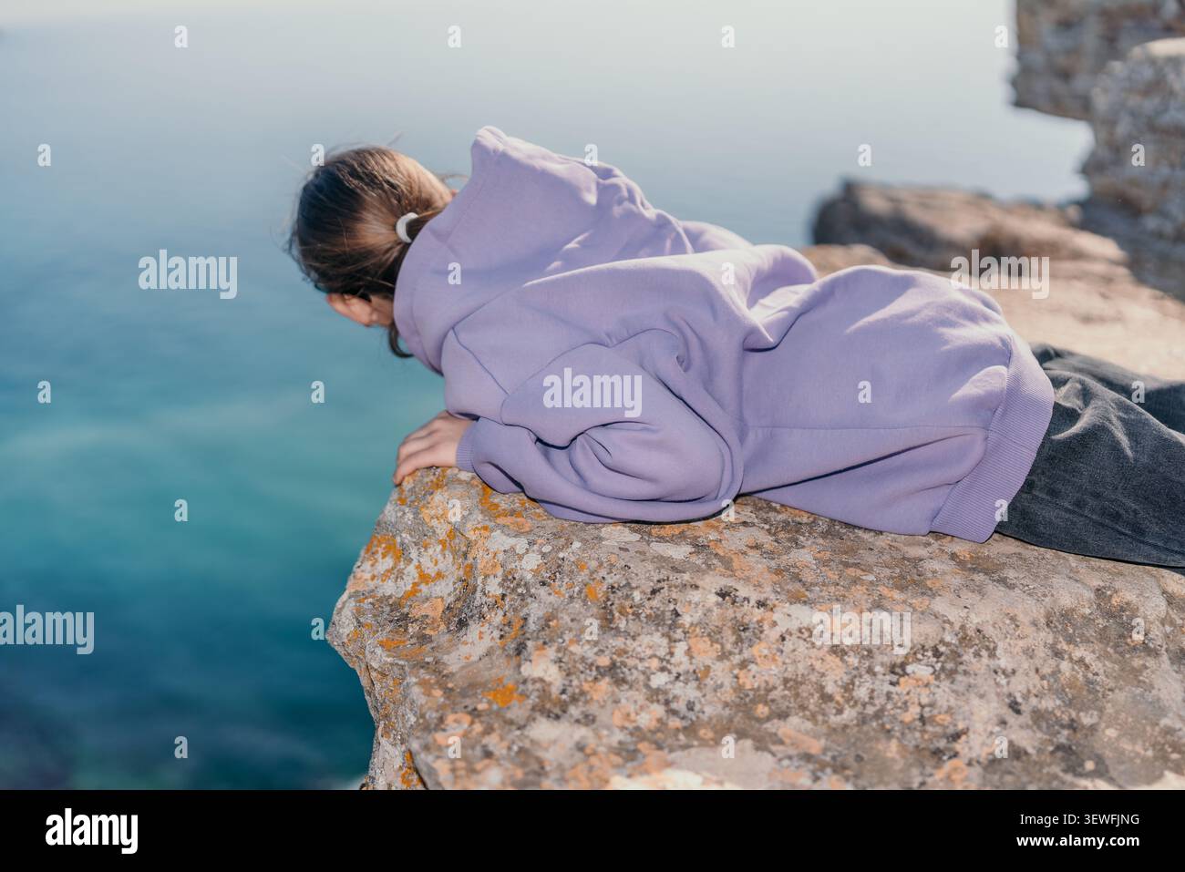 Blick auf das Meer einer Kinderklippe auf ein Mädchen, das auf einem felsigen Bergrücken liegt und in tiefblaues Wasser hinunterblickt Stockfoto