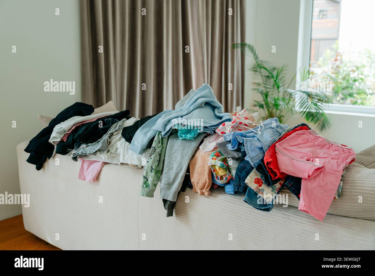 Ein großer, unordentlicher Stapel bunter Babykleidung und Kleidungsstücke, die auf einem hellen Sofa im Wohnzimmer gestapelt sind und darauf warten, gefaltet oder gebügelt zu werden. Stock Foto Stockfoto