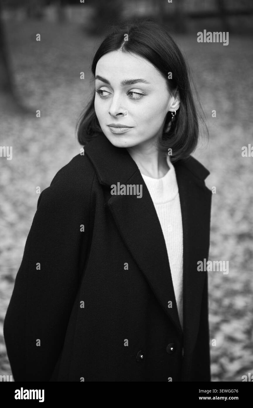 Einfarbiges Porträt einer jungen Frau im dunklen Mantel, die im Herbstpark zur Seite blickt, eingerahmt von weichem Laub. Ideal für Mode, eleganten Lifestyle, zeitloses Branding und Werbung Stockfoto