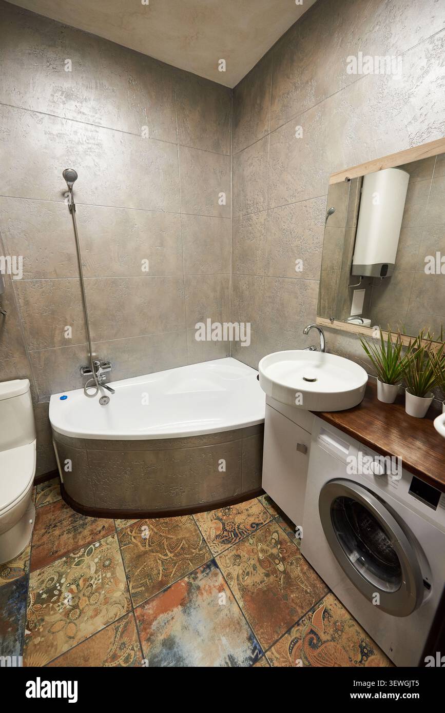 Modernes Bad mit eingebauter Badewanne und Waschbecken auf Holzwaschtisch. Kompaktes Badezimmer mit Steinfliesen, Spiegel, Waschmaschine, Handtuchhalter und Topfpflanzen Stockfoto