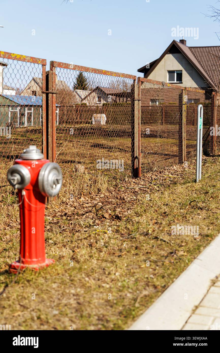 Ein knallroter, klassischer Feuerhydrant steht an einer grasbewachsenen Straße neben einem Drahtzaun in einer ruhigen Wohngegend. Stockfoto