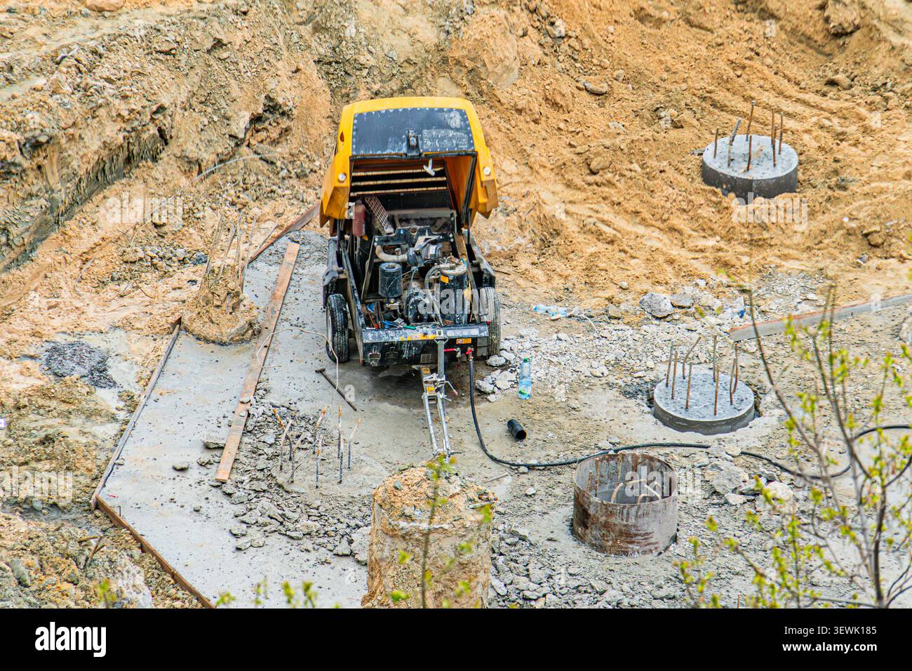 Dynamische Bauszene mit mobiler Betonpumpe, zylindrischen Formen, Bewehrung, ausgehobener Erde, teilweise gegossenem Beton, grüne Pflanzen, hell Stockfoto