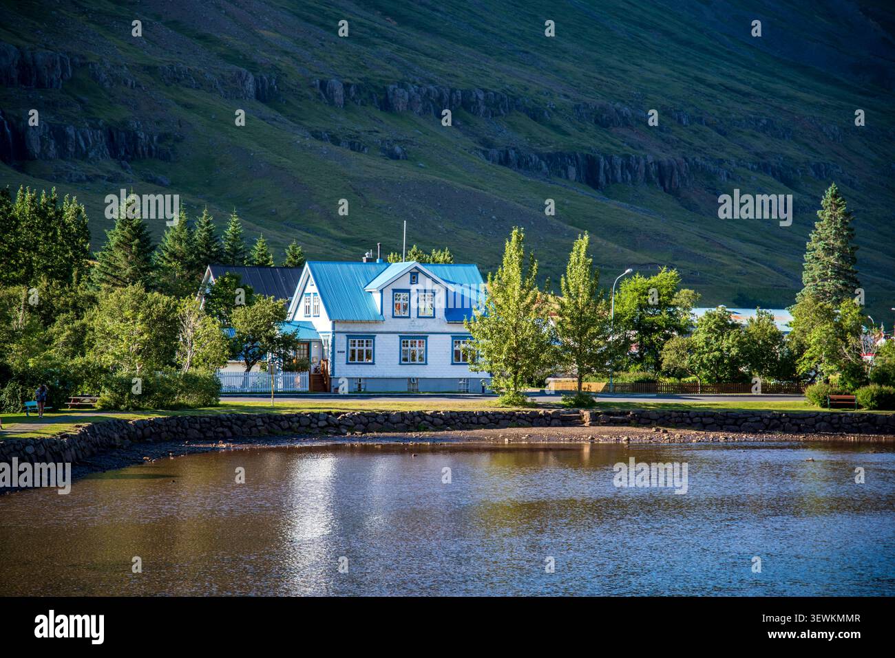 Wunderschönes traditionelles Haus mit einem hellblauen Dach, das entlang der malerischen Uferpromenade unter steilen grünen Bergen in Seyðisfjörður, Ost-Island, liegt. Stockfoto