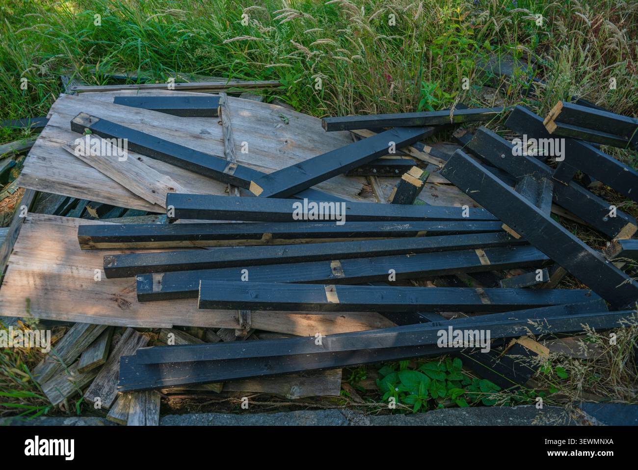 Verschiedene Holzabfälle, darunter mehrere dunkel lackierte Balken, die auf verwitterten Brettern zwischen grüner Vegetation und hohem Gras ruhen Stockfoto