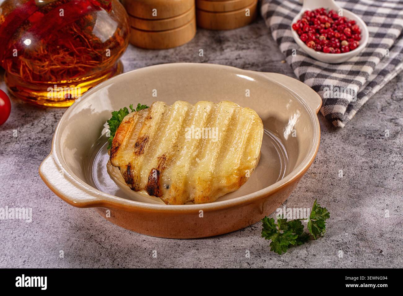 Gegrillte halloumi-Käsescheibe mit goldenen Grillspuren in Keramikschale. Frische Petersilie, Safranöl und rosa Pfeffer auf grauem Steinhintergrund Stockfoto