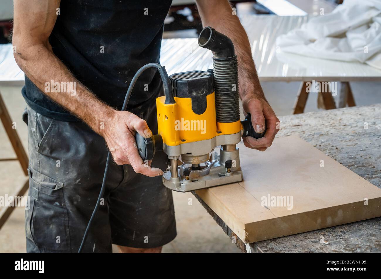 Ein Zimmermann betreibt in einer Holzwerkstatt einen gelben Tauchfräser auf einer Holzplatte und formt Kanten und Oberflächen mit präzisen Elektrowerkzeugen Stockfoto