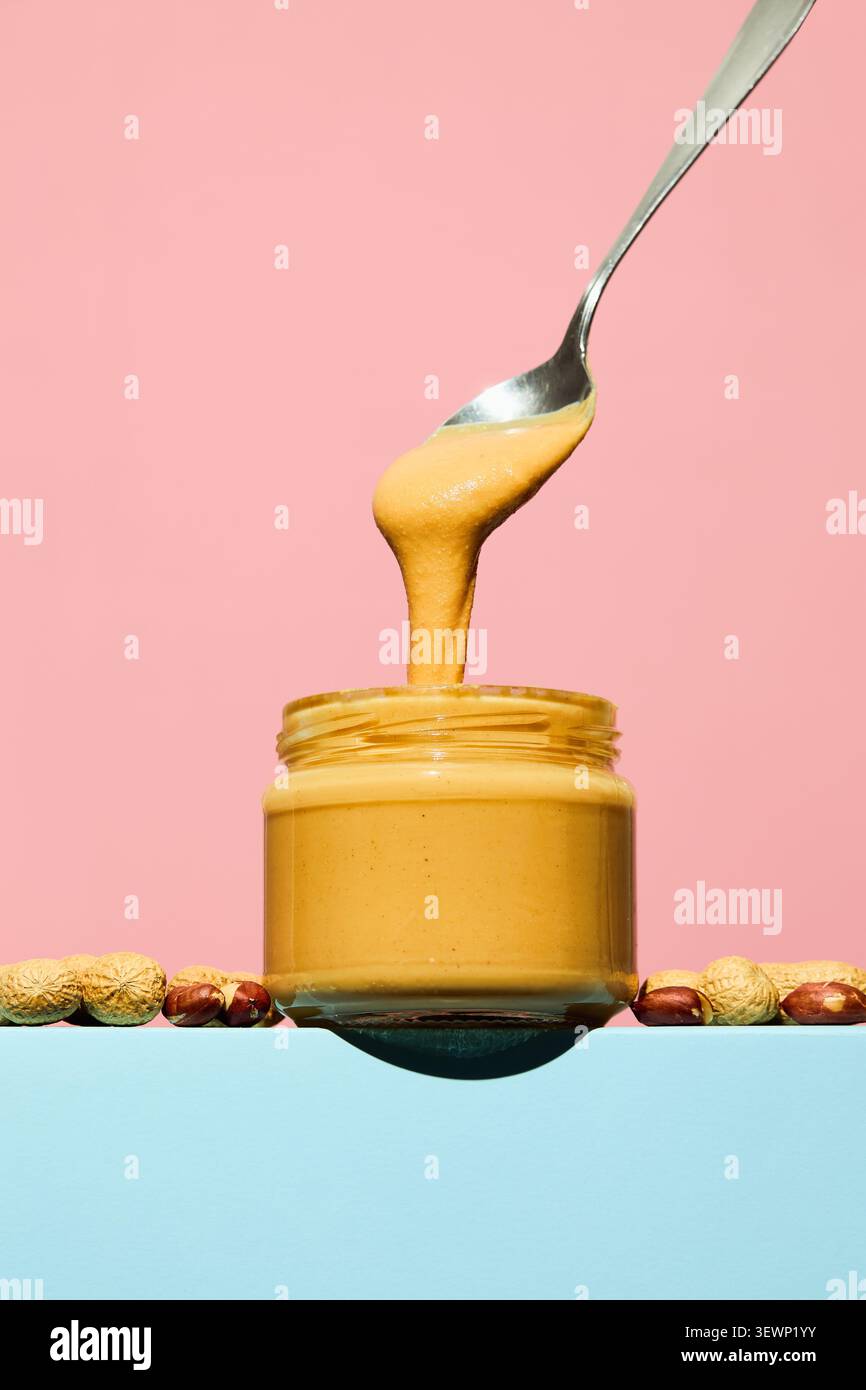 Cremige Erdnussbutter, die aus einem Löffel in ein Glas mit verstreuten Erdnüssen auf pastellfarbenem Hintergrund fließt. Stockfoto