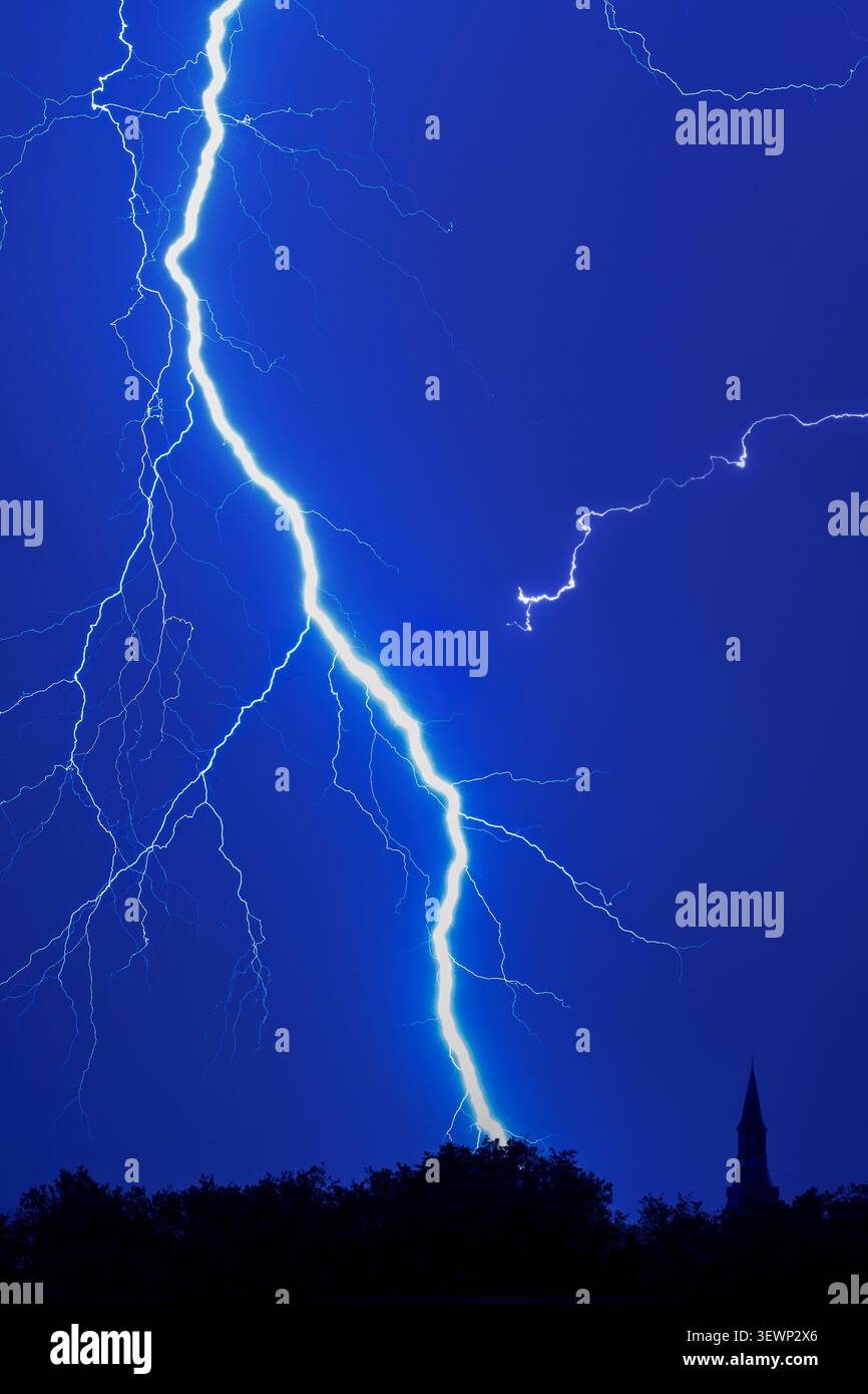 Gespaltener Blitz / Wolken-zu-Boden-Blitzschlag während eines Gewitters am blauen Nachthimmel über Wald mit Silhouettenbaumkronen und Kirchturm, Stockfoto