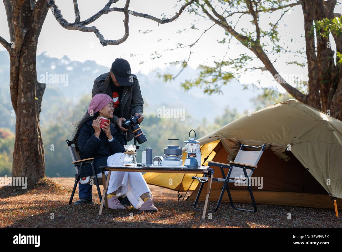 Ein Paar entspannt sich auf einem Campingplatz mit Ausrüstung, während es sich die Bilder der Digitalkamera in einer ruhigen Umgebung im Freien ansieht. Stockfoto