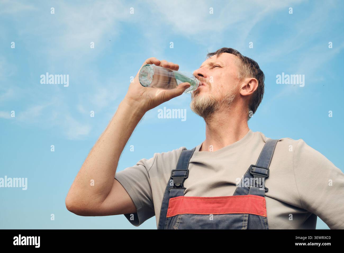 Mann im Overall, der während der Arbeitspause mit blauem Himmel hydriert Stockfoto