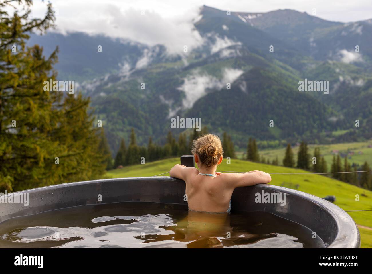 Frau, die sich im Außenpool mit Blick auf die Berge entspannt. Rückansicht einer blonden Frau, die in einer Badewanne im Freien mit Blick auf grüne, immergrüne Bergwiesen taucht Stockfoto