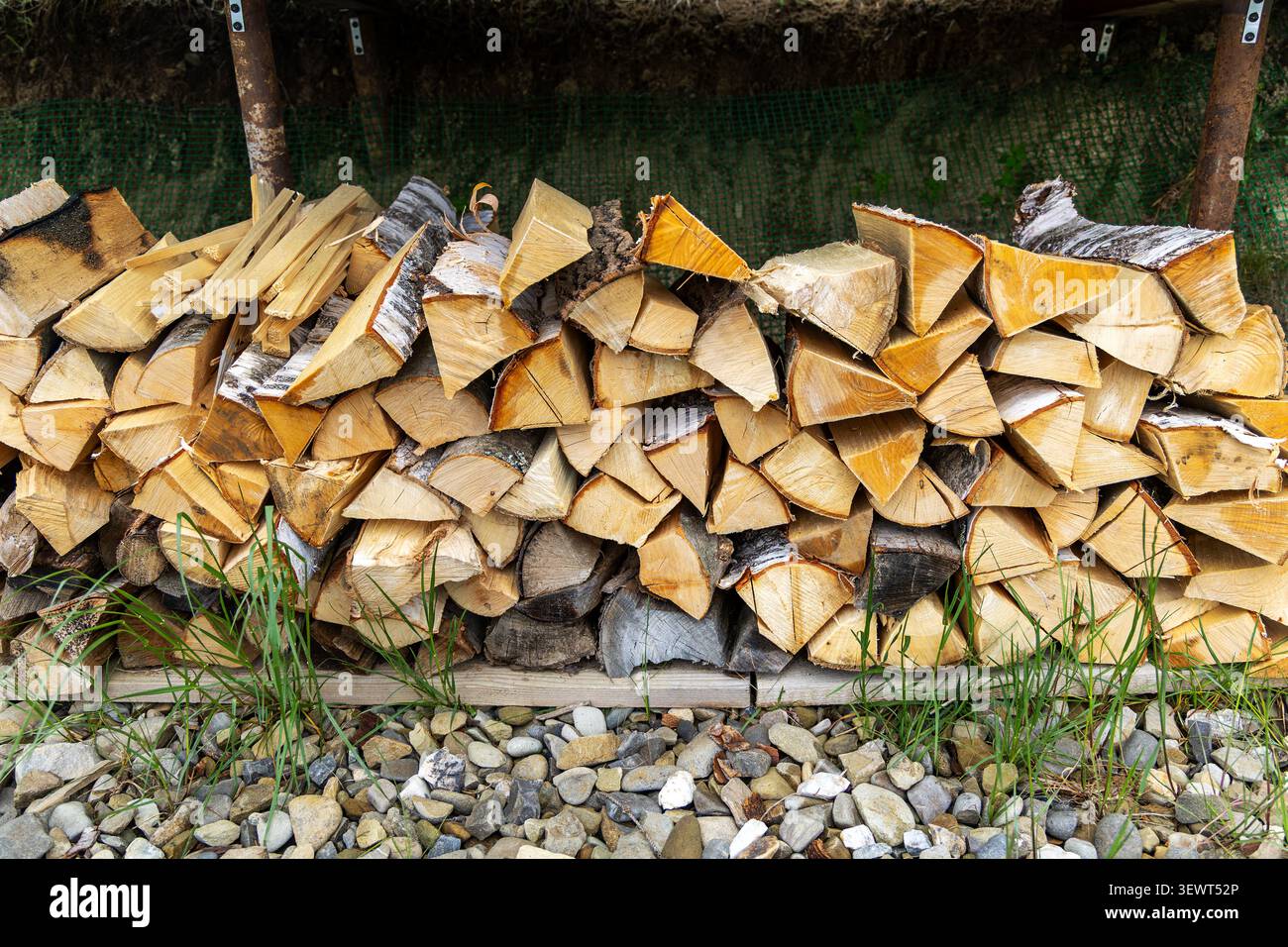 Gestapeltes Feuerholz Unter Einem Dach Im Freien. Gestapeltes Brennholz unter einem Außenbereich mit Birkenrinde, rauen Holzflächen, grünem Gras und Kies Stockfoto
