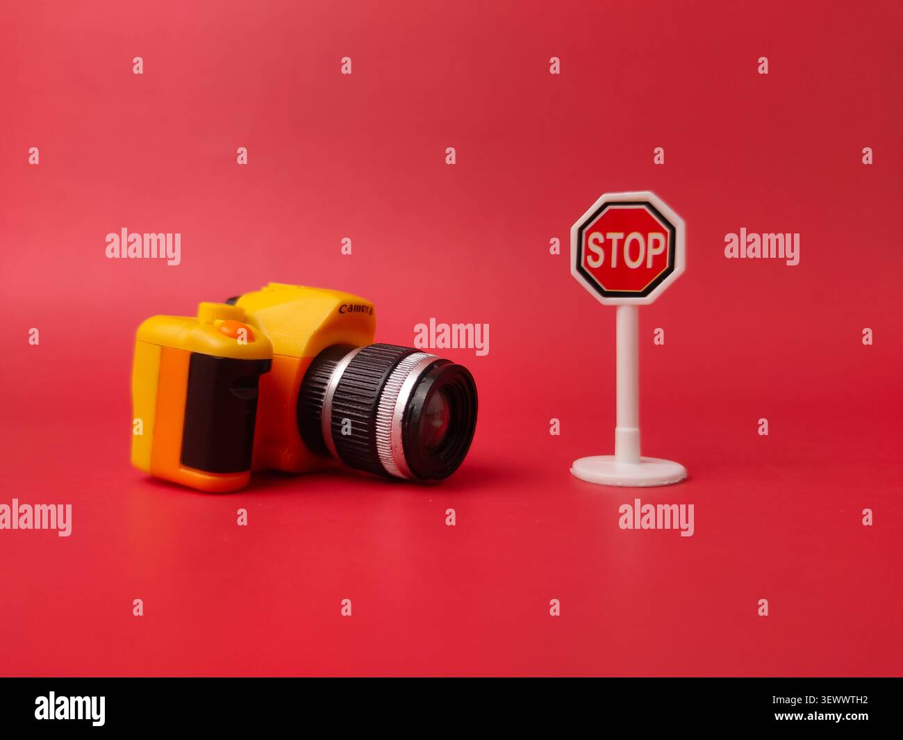 Eine gelbe Miniatur-Spielzeugkamera liegt flach neben einem kleinen weißen achteckigen Stoppschild auf einem durchgehend roten Hintergrund. Stockfoto