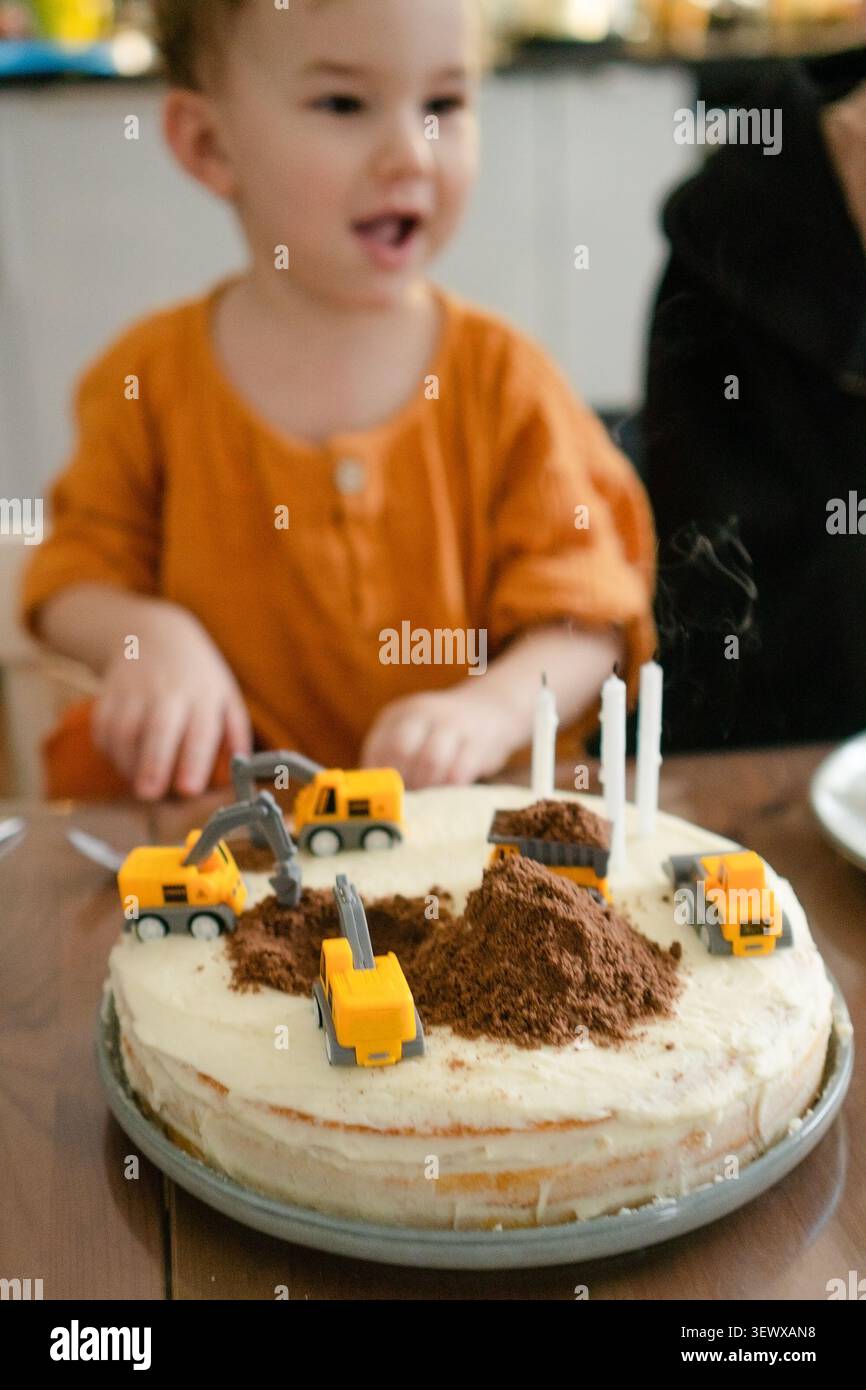Der kleine Junge lächelt, nachdem er erfolgreich die Kerzen auf seiner Geburtstagskuche ausgeblasen hat. Das Kind ist drei Jahre alt. Hausgemachter Kuchen ist wie eine Baustelle mit Spielzeugwagen dekoriert. Familienfeier Stockfoto