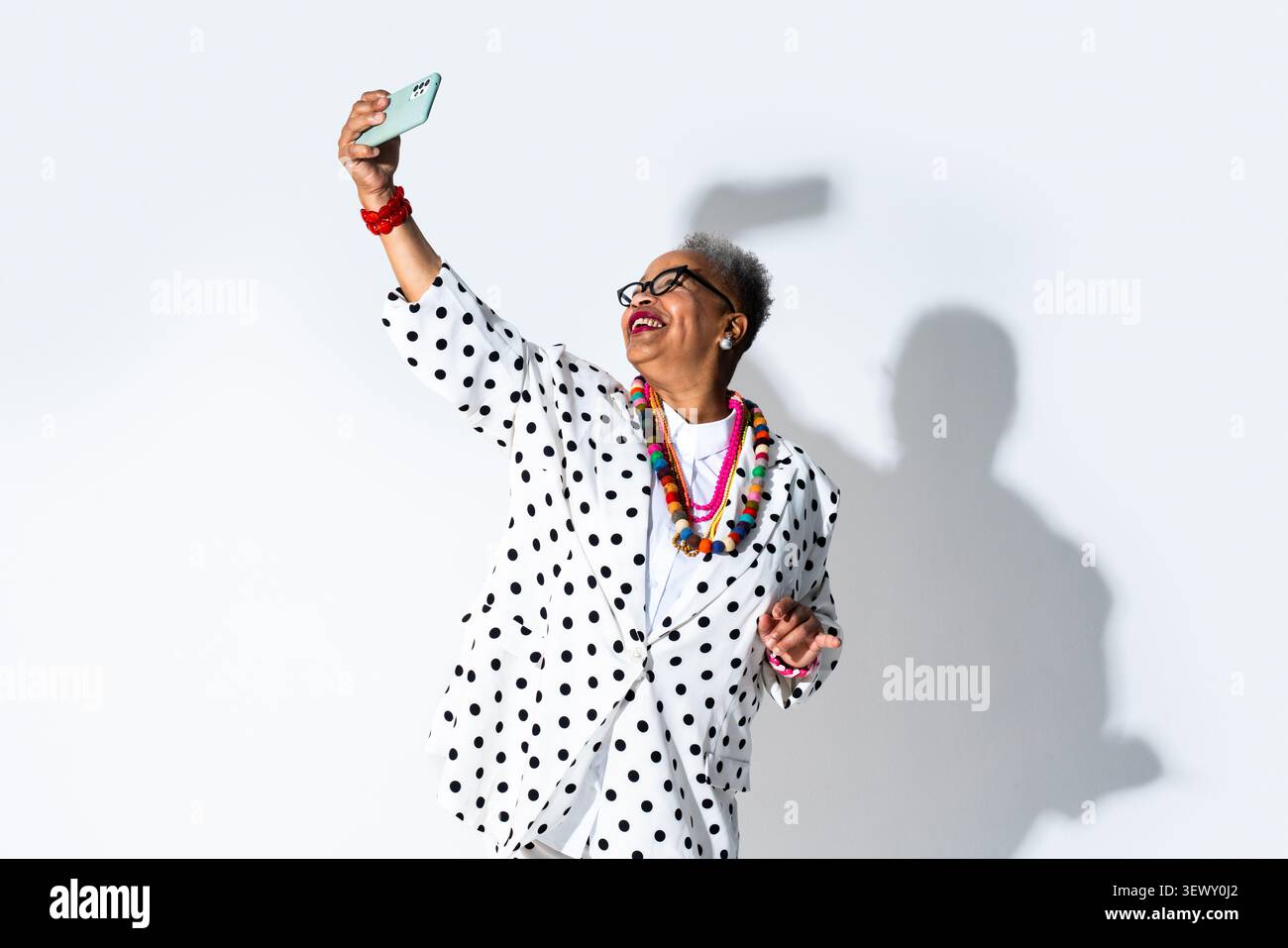 Schwarze Seniorin trägt moderne Kleidung Porträt auf Hintergrund - Seniorin mit kurzen grauen Haaren und Brille, die ein Selfie mit ihrem Smartphone macht, Stockfoto