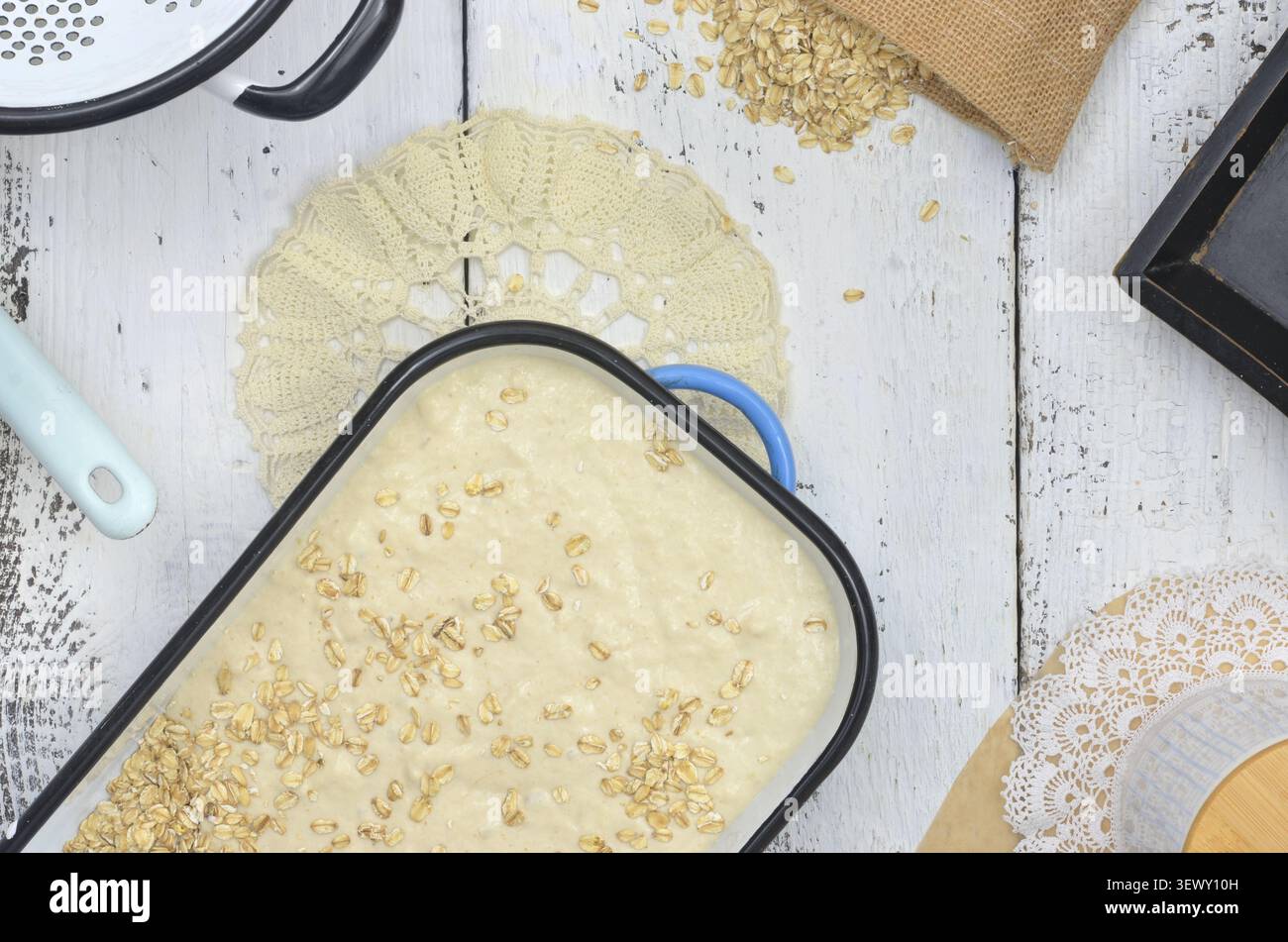 Gerösteter, roher Hefeteig, bestreut mit Haferflocken in blauer, emaillierter Kuchenform, bereit zum Backen. Emaillierte Küchenutensilien auf einem Holzbalkon. Stockfoto
