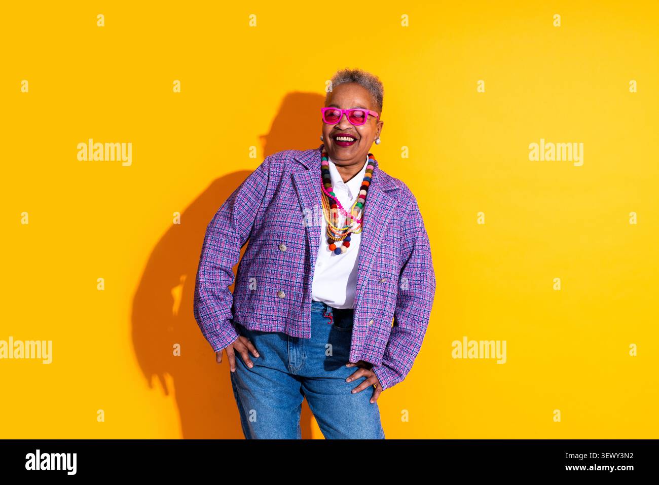 Schwarze ältere Frau trägt moderne Kleidung Porträt auf Hintergrund - ältere afroamerikanische Frau lacht. Sie trägt eine bunte Brille und ein schickes Outfit Stockfoto